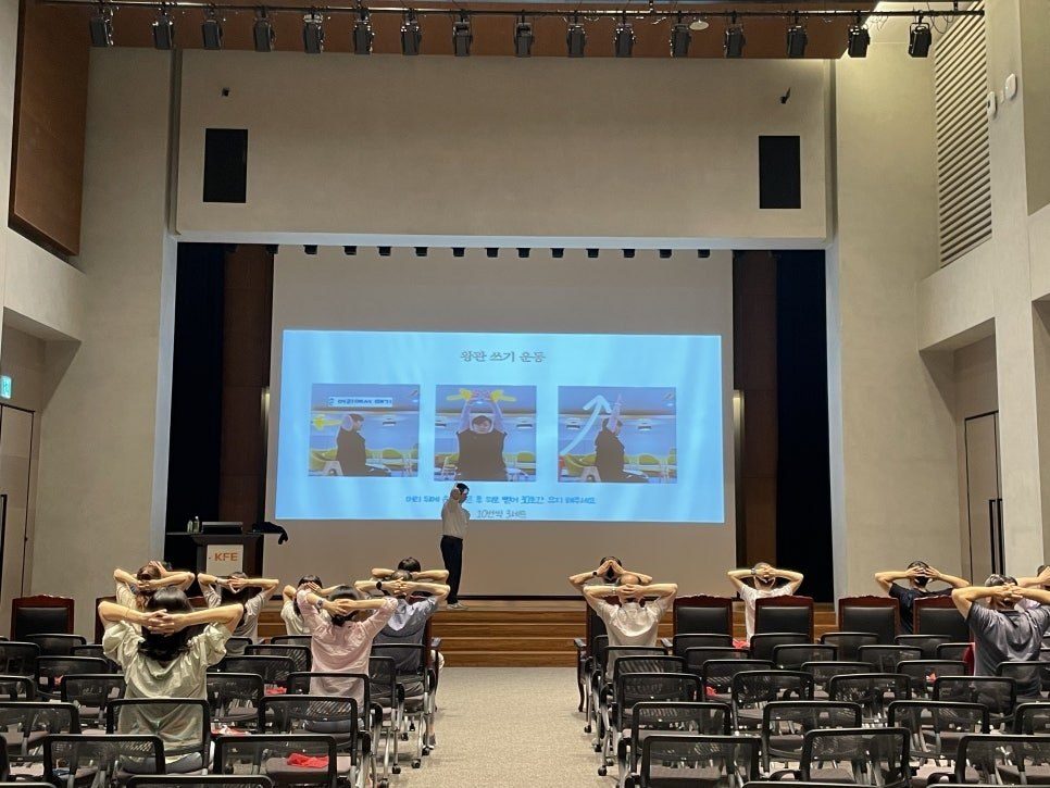 대강당 객석의 임직원들이 두 손을 머리 뒤에 두고 동작을 따라 하는 뒷모습